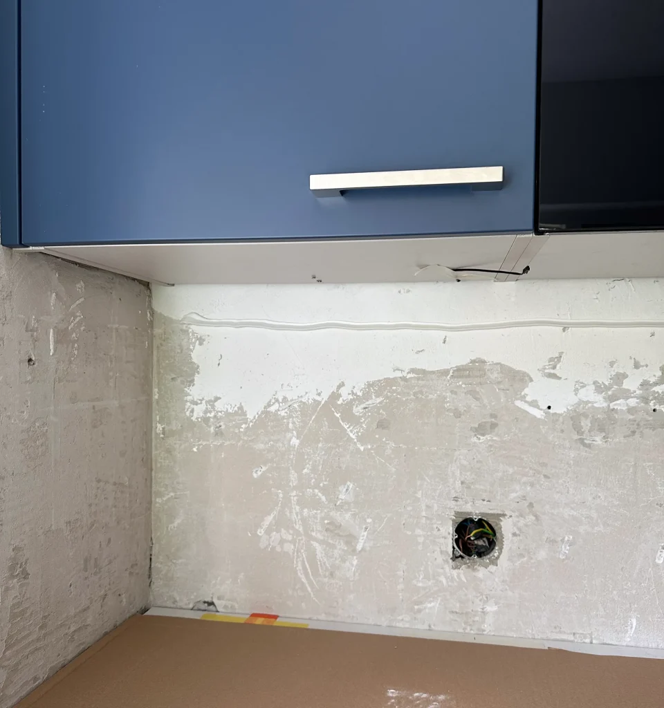 Bare kitchen wall under the wall cabinets, ready for the backsplash, with the socket cover removed and the worktop covered with cardboard